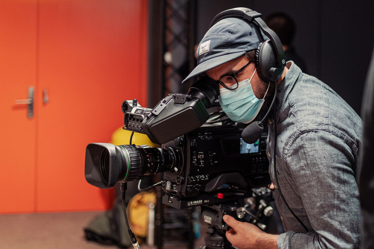 Ein Kameramann mit Maske und Kopfhörern bedient eine professionelle Videokamera vor einer orangefarbenen Tür.