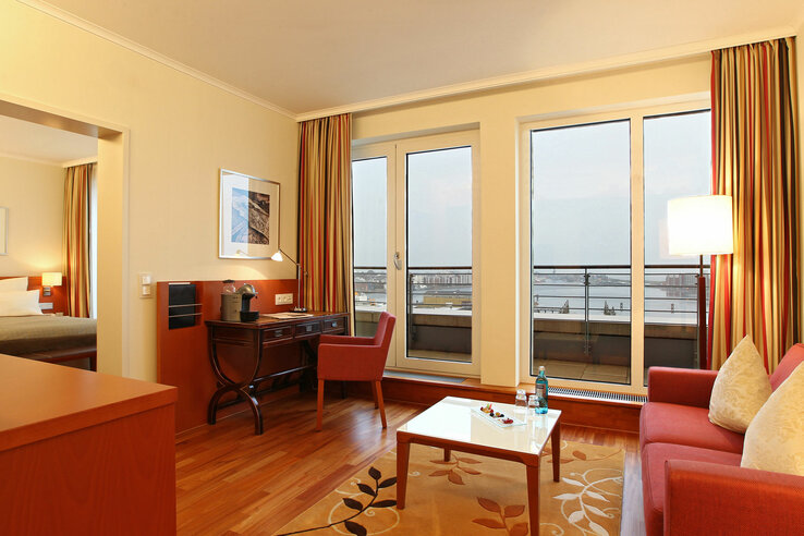 Helles Hotelzimmer mit Sofa, Schreibtisch, großen Fenstern mit Stadtblick und Zugang zu einem angrenzenden Schlafzimmer.