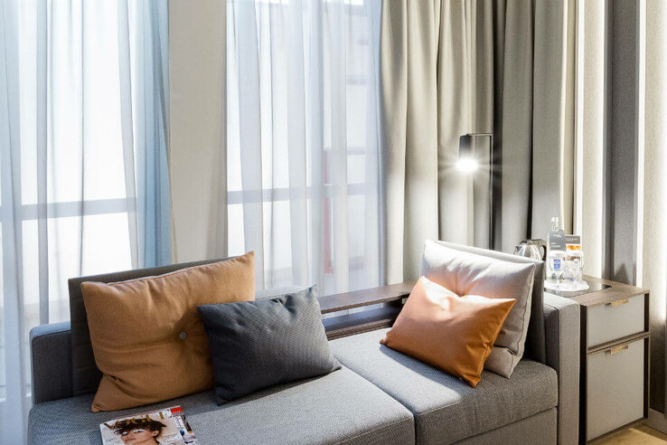 Bright hotel room with grey sofa, colorful cushions, side table, lamp and windows with curtains.