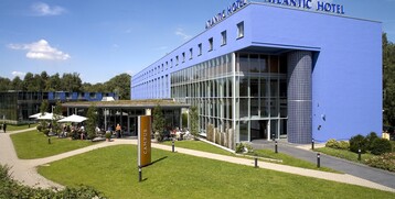 ATLANTIC Hotel Universum in Bremen, modern blue building with glass facade, green surroundings, outdoor terrace with guests.