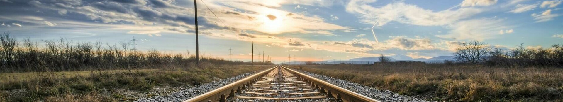 Mit der deutschen Bahn zu den ATLANTIC Hotels reisen Gleise führen in den Horizont, umgeben von Feldern, unter einem dramatischen Himmel bei Sonnenuntergang.