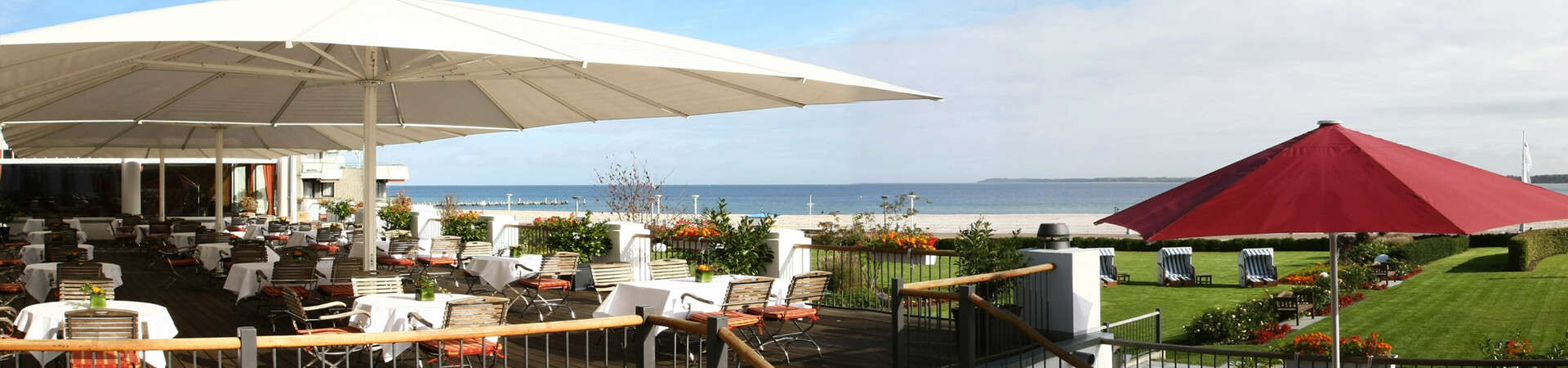Terrace of the ATLANTIC Grand Hotel Travemünde with sea view, tables, chairs and sunshades, surrounded by garden.