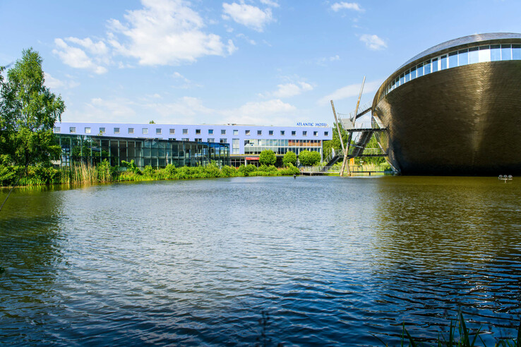 ATLANTIC Hotel Universum in Bremen, modernes Gebäude am See, blauer Himmel und grüne Vegetation im Vordergrund.