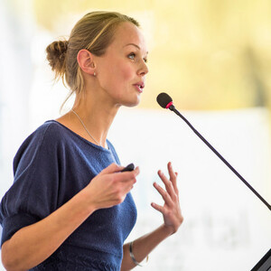 Eine Frau hält eine Präsentation mit Mikrofon, trägt ein blaues Oberteil und hat blondes Haar.