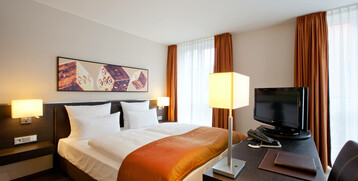 Modernes Hotelzimmer mit Doppelbett, orangefarbenem Überwurf, Schreibtisch, Flachbild-TV und großen Fenstern mit Vorhängen.