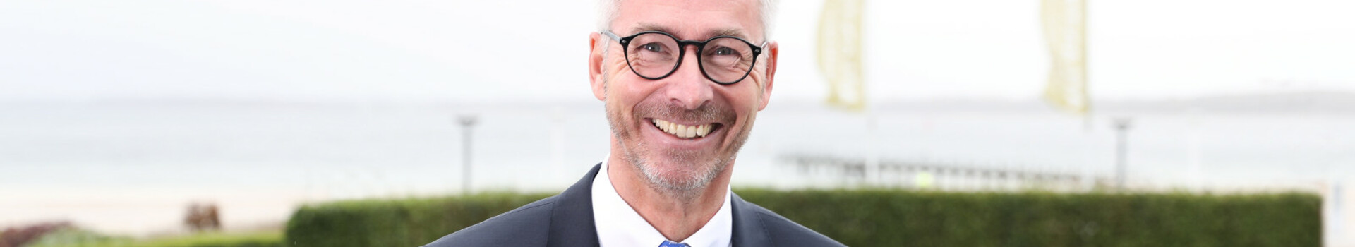 Ein lächelnder Mann in Anzug vor einer Küstenlandschaft mit Meer und Grünflächen im Hintergrund.