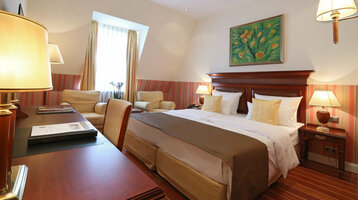 Gemütliches Hotelzimmer mit Doppelbett, Holzmöbeln, Sesseln, warmem Licht und Blumenbild an der Wand.