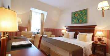 Gemütliches Hotelzimmer mit Doppelbett, Holzmöbeln, Sesseln, warmem Licht und Blumenbild an der Wand.