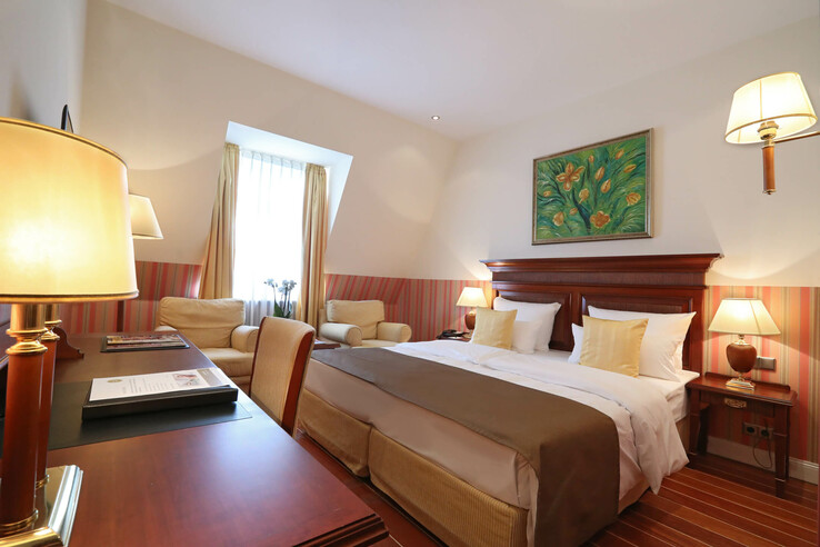 Gemütliches Hotelzimmer mit Doppelbett, Holzmöbeln, Sesseln, warmem Licht und Blumenbild an der Wand.