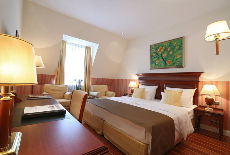 Gemütliches Hotelzimmer mit Doppelbett, Holzmöbeln, Sesseln, warmem Licht und Blumenbild an der Wand.