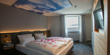 Modernes Hotelzimmer mit Doppelbett, Rosenblättern, Himmel-Decke, Fenster und TV im ATLANTIC Hotel Airport Bremen.