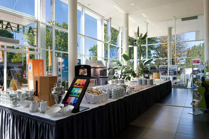 Catering in der Lobby vom ATLANTIC Hotel Galopprennbahn Helles Frühstücksbuffet mit Gebäck, Tee und Geschirr im ATLANTIC Hotel Galopprennbahn, umgeben von großen Fenstern und Pflanzen.
