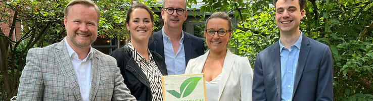 Fünf Personen lächeln in einem Garten, halten ein "Greensign Hotel"-Schild.