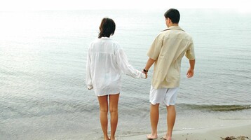 Couple holding hands and standing barefoot on a quiet beach overlooking the sea.