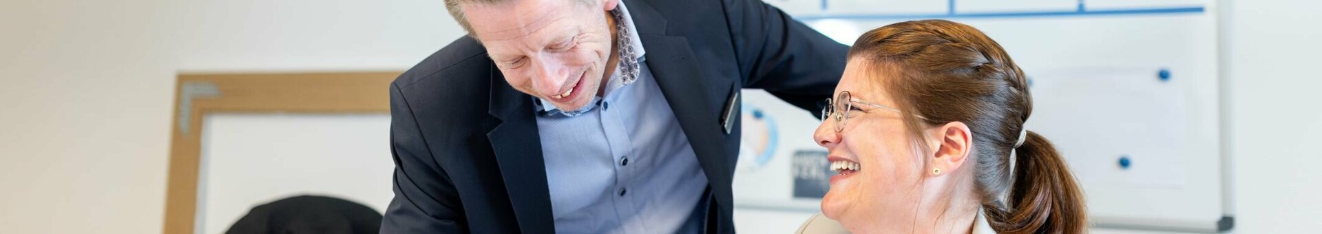 Zwei Personen in Business-Kleidung lachen gemeinsam in einem Büro vor einem Whiteboard.