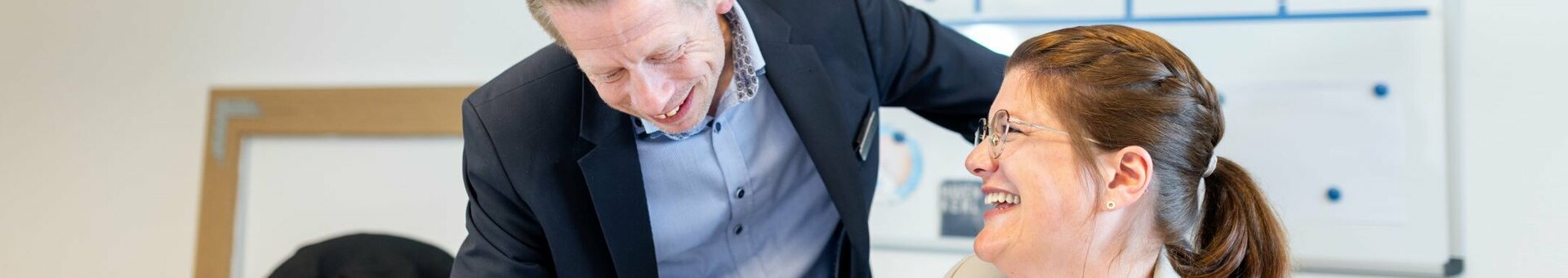 Two people in business attire laughing together in an office in front of a whiteboard.