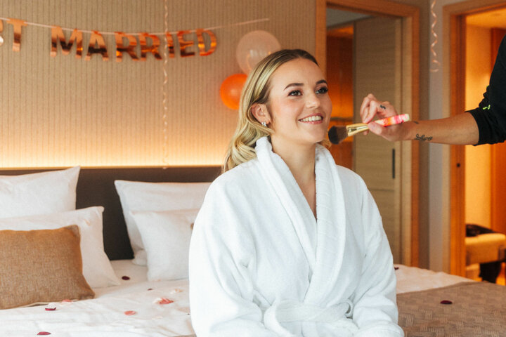 Woman in bathrobe being made up in hotel room, "Just Married" decoration in the background.