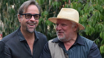 Zwei Männer lachen im Freien; einer hält ein Handy mit ATLANTIC Hotel Münster-Hülle.