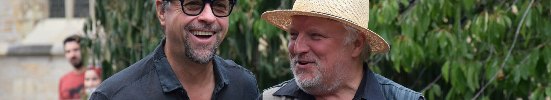 ATLANTIC Hotel Münster | Angebot KRIMI & TATORTSTADT MÜNSTER Zwei Männer lachen im Freien; einer hält ein Handy mit ATLANTIC Hotel Münster-Hülle.