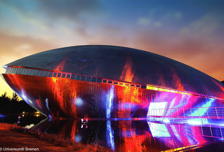 Futuristisches Gebäude in Bremen, beleuchtet in bunten Farben bei Sonnenuntergang, spiegelt sich in Wasserfläche.