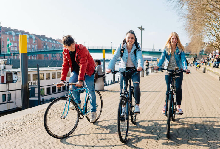 Drei Personen fahren bei sonnigem Wetter auf Fahrrädern entlang einer Uferpromenade, im Hintergrund sind Boote und Gebäude.