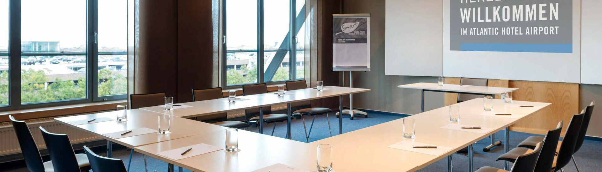Veranstaltungsraum Dornier im ATLANTIC Hotel Airport Bremen Konferenzraum mit U-förmiger Bestuhlung, Fenstern, Flipchart und Begrüßungstext im ATLANTIC Hotel Airport Bremen.