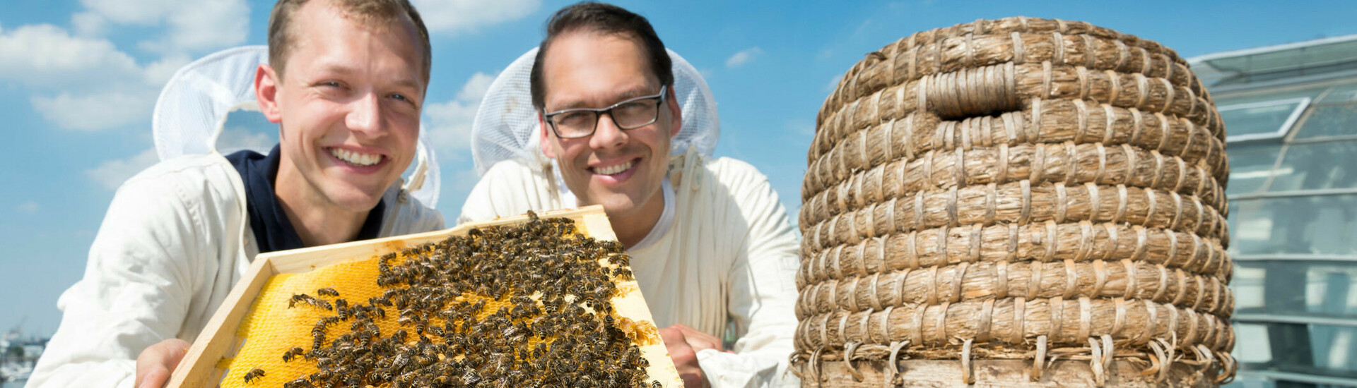 Two beekeepers show a honeycomb next to a traditional beehive on the roof of the ATLANTIC Hotel Sail City.