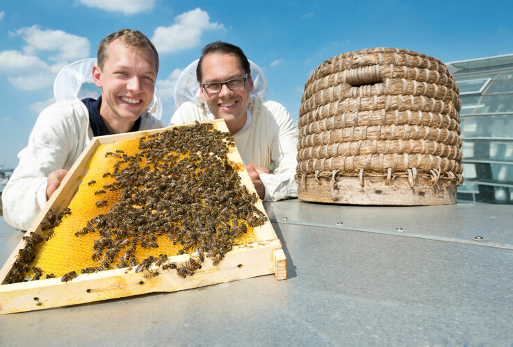 Zwei Imker zeigen eine Bienenwabe neben einem traditionellen Bienenkorb auf dem Dach des ATLANTIC Hotel Sail City.