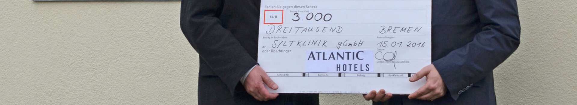 Syltklinik: Erholung für Familien mit einem krebskranken Kind Zwei Männer überreichen einen Scheck über 3.000 Euro vor einem bunten Schild der Sylt Klinik.