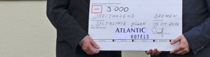 Syltklinik: Erholung für Familien mit einem krebskranken Kind Zwei Männer überreichen einen Scheck über 3.000 Euro vor einem bunten Schild der Sylt Klinik.
