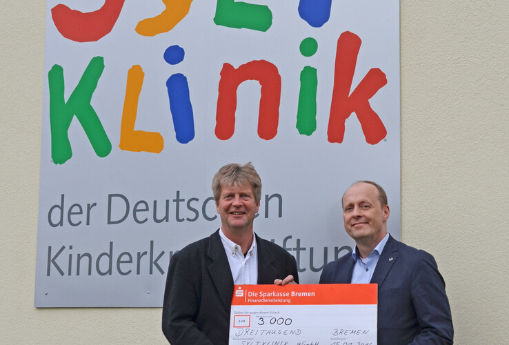 Syltklinik: Erholung für Familien mit einem krebskranken Kind Zwei Männer überreichen einen Scheck über 3.000 Euro vor einem bunten Schild der Sylt Klinik.