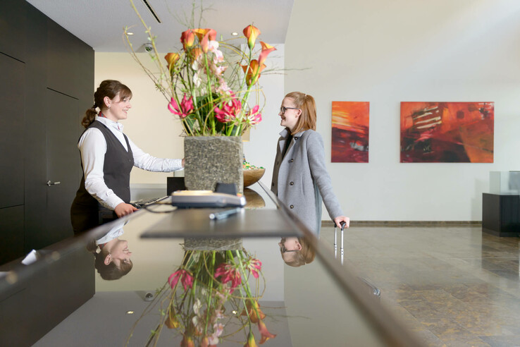 Hotelrezeption mit Blumenarrangement, Empfangsdame lächelt Gast mit Koffer an, moderne Kunst im Hintergrund.