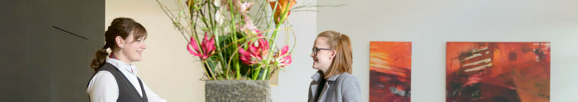 ATLANTIC Hotel Lübeck – Empfang Hotelrezeption mit Blumenarrangement, Empfangsdame lächelt Gast mit Koffer an, moderne Kunst im Hintergrund.