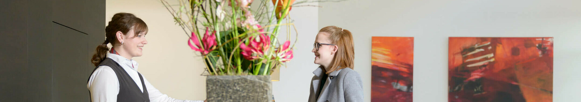 Hotel reception with flower arrangement, receptionist smiles at guest with suitcase, modern art in the background.