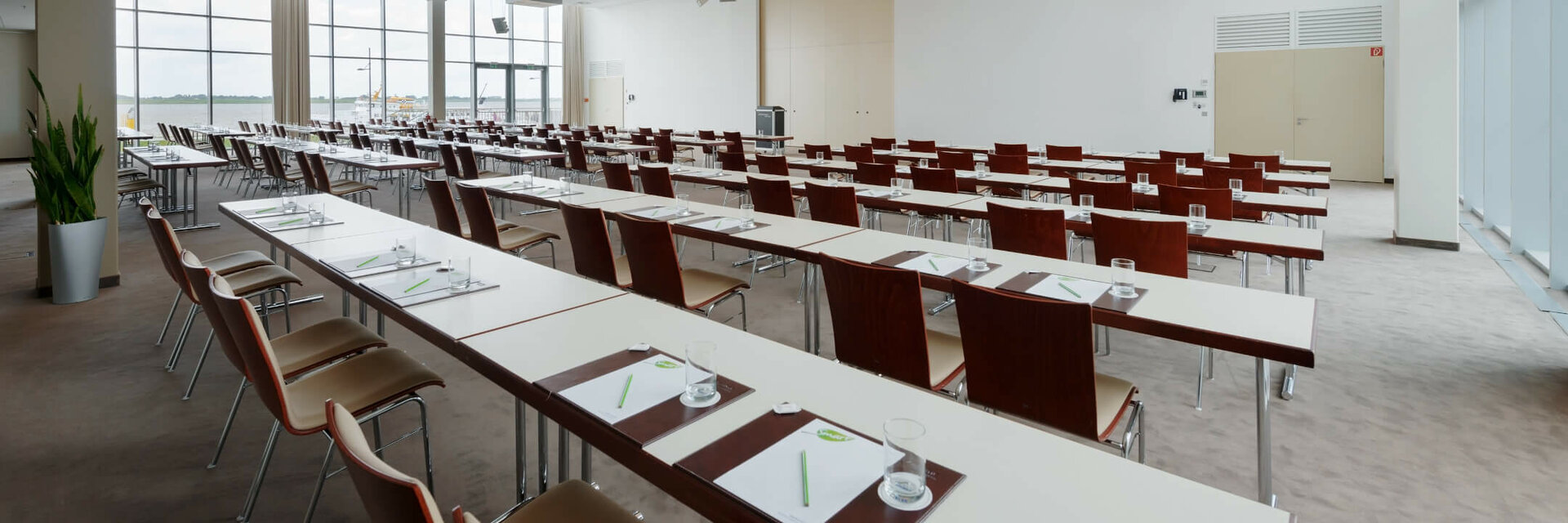 Moderner, heller Konferenzraum mit Reihen aus Tischen und Stühlen, großen Fenstern und Schreibmaterialien.