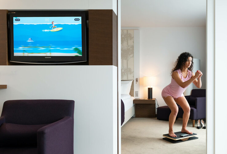 Woman doing fitness exercises on balance board in modern hotel room, TV shows surfing simulation.
