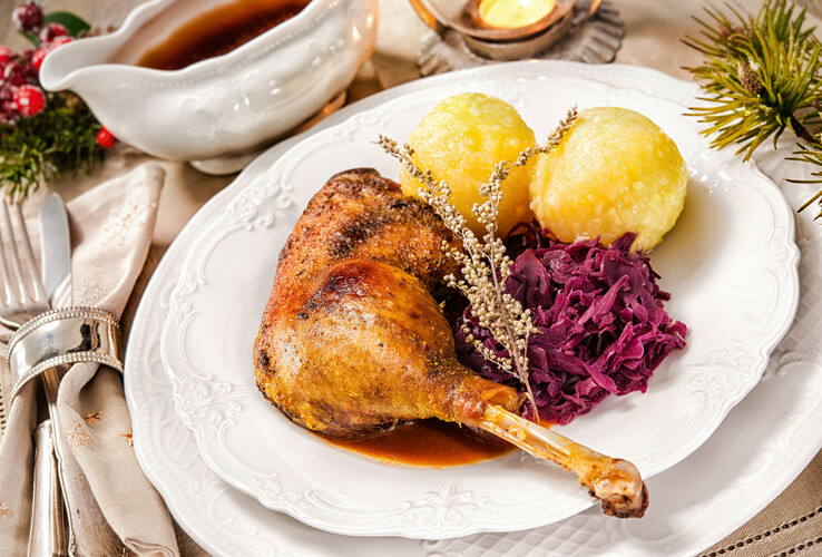 Festive dish with duck leg, red cabbage and potato dumplings on a white plate, decorated with herbs.