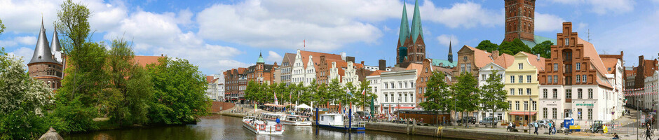 Historische Altstadt von Lübeck mit Backsteinhäusern am Wasser, blauer Himmel und grüne Bäume.