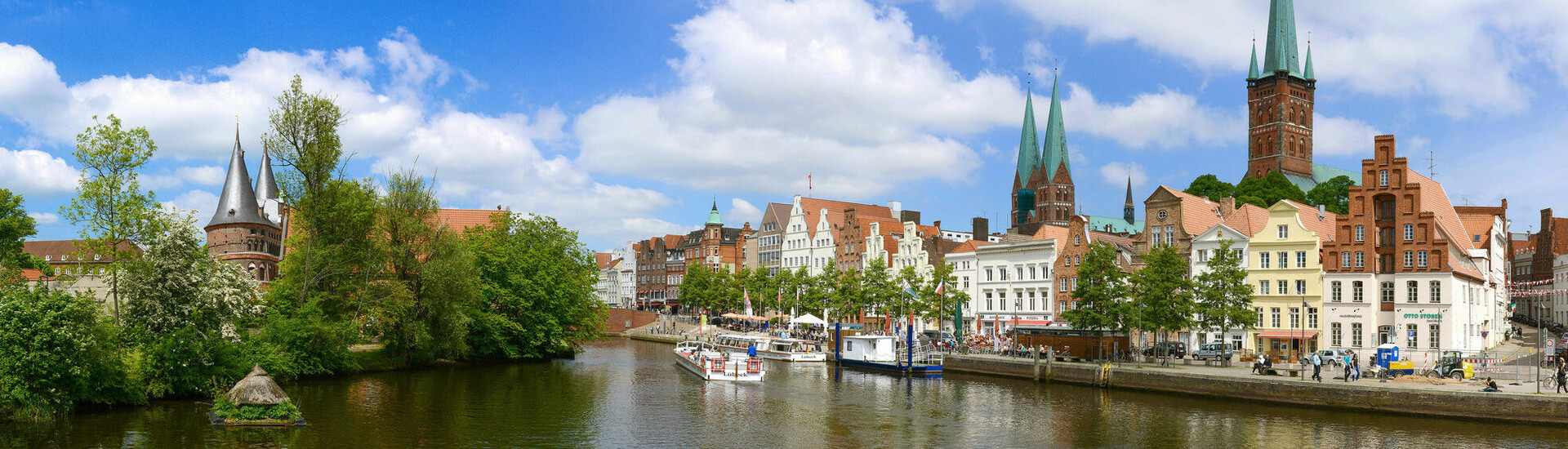 Blick auf die Lübecks Altstadt an der Untertrave Historische Altstadt von Lübeck mit Backsteinhäusern am Wasser, blauer Himmel und grüne Bäume.