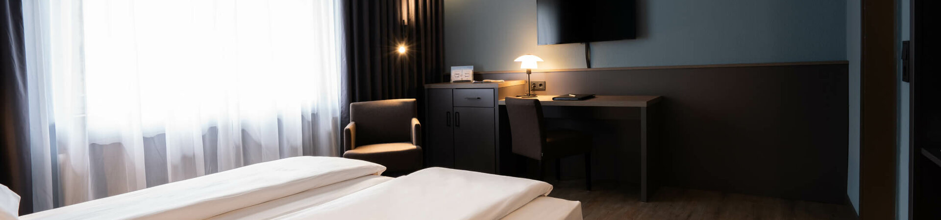 Modernes Hotelzimmer mit Doppelbett, Schreibtisch, Fernseher, Sessel und großen Fenstern mit Vorhängen.