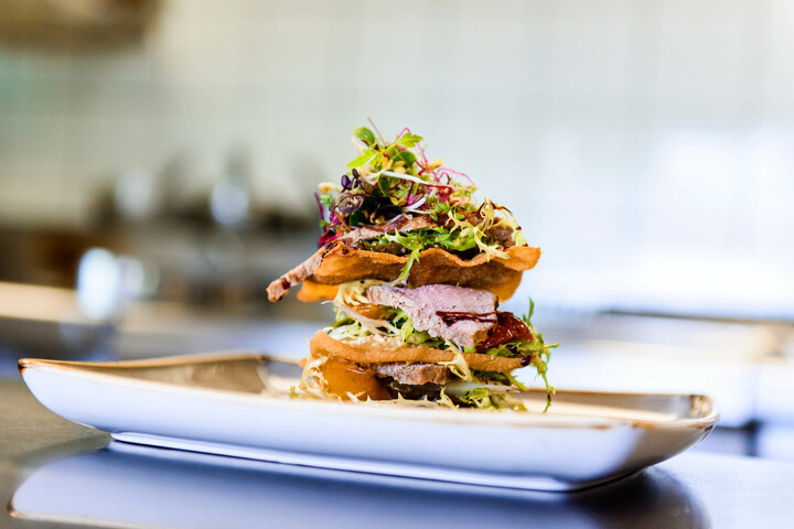 Speisen für Tagungen und Feiern im ATLANTIC Hotel Universum Fein angerichtetes Gourmetgericht mit Fleisch, Salat und knusprigen Schichten auf einem eleganten Teller.