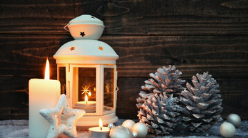 Weiße Laterne mit Kerzen, Tannenzapfen und Weihnachtskugeln auf verschneitem Holzuntergrund. Stimmungsvolle Winterdekoration.