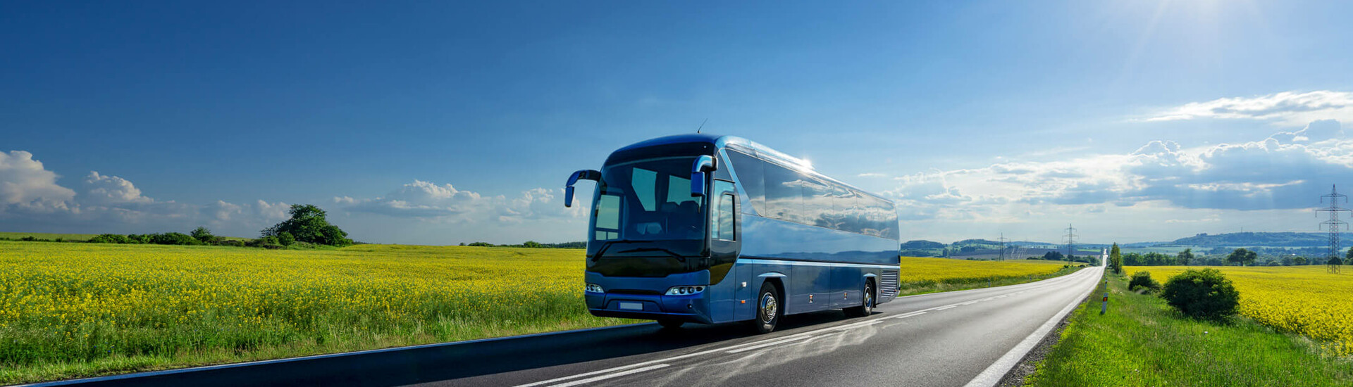 Coach on a country road, surrounded by yellow fields under a blue sky with sun.