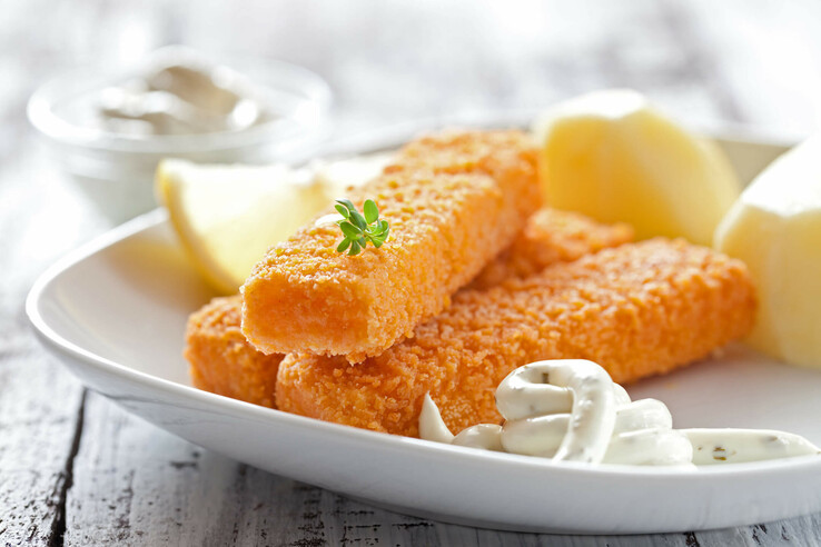 Panierte Fischstäbchen mit Zitronenscheibe, Kartoffeln und Kräuterdip auf einem weißen Teller.