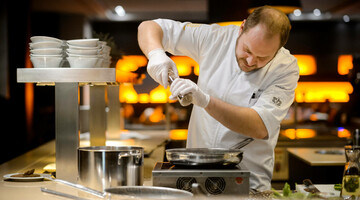 Koch in weißer Uniform würzt ein Gericht in einer modernen Hotelküche.