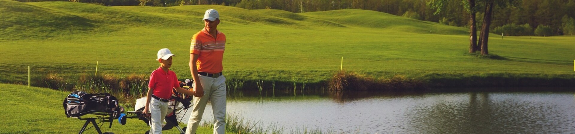 Zwei Personen beim Golfen auf grünem Platz neben Teich, unter blauem Himmel mit Wolken.