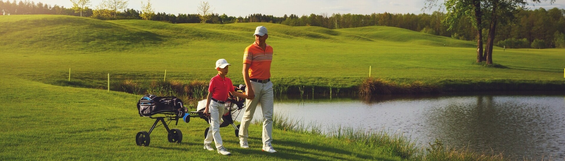 Two people playing golf on a green course next to a pond, under a blue sky with clouds.