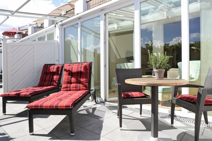 Sonnige Hotelterrasse mit zwei roten Liegen, Tisch, Stühlen und Pflanze vor verglaster Fassade.