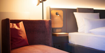 Modernes Hotelzimmer mit gemütlichem Sessel, Kissen, Bett und stilvoller Beleuchtung im ATLANTIC Hotel Airport Bremen.