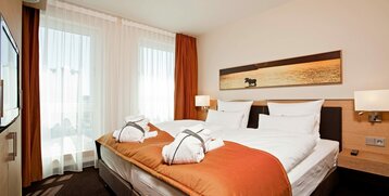 Modernes Hotelzimmer mit Doppelbett, weißen Bettlaken, orangefarbener Decke und Bademänteln, helle Vorhänge.